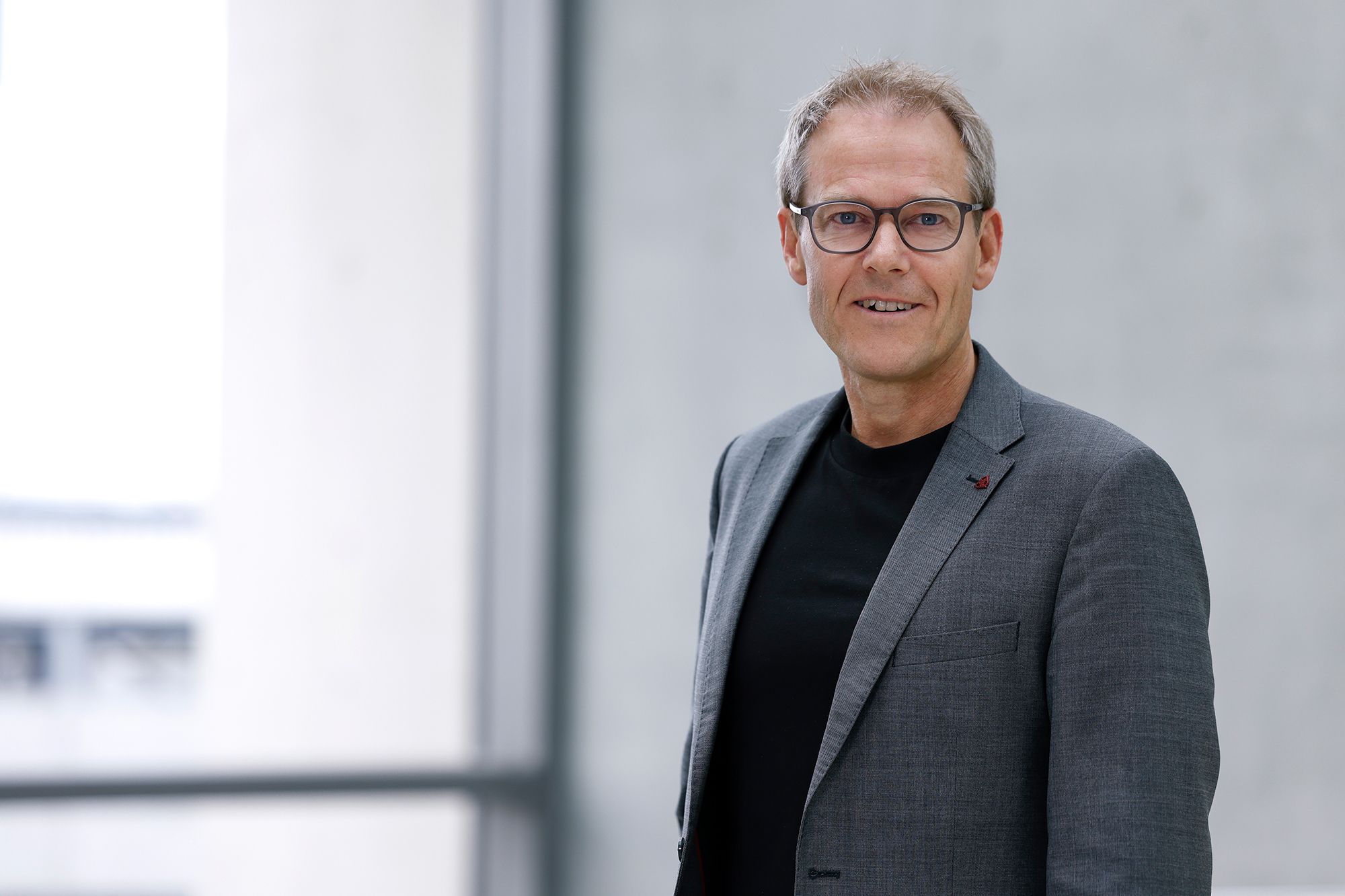 Mann mit Brille und grauen Anzug vor einer Fensterfront.