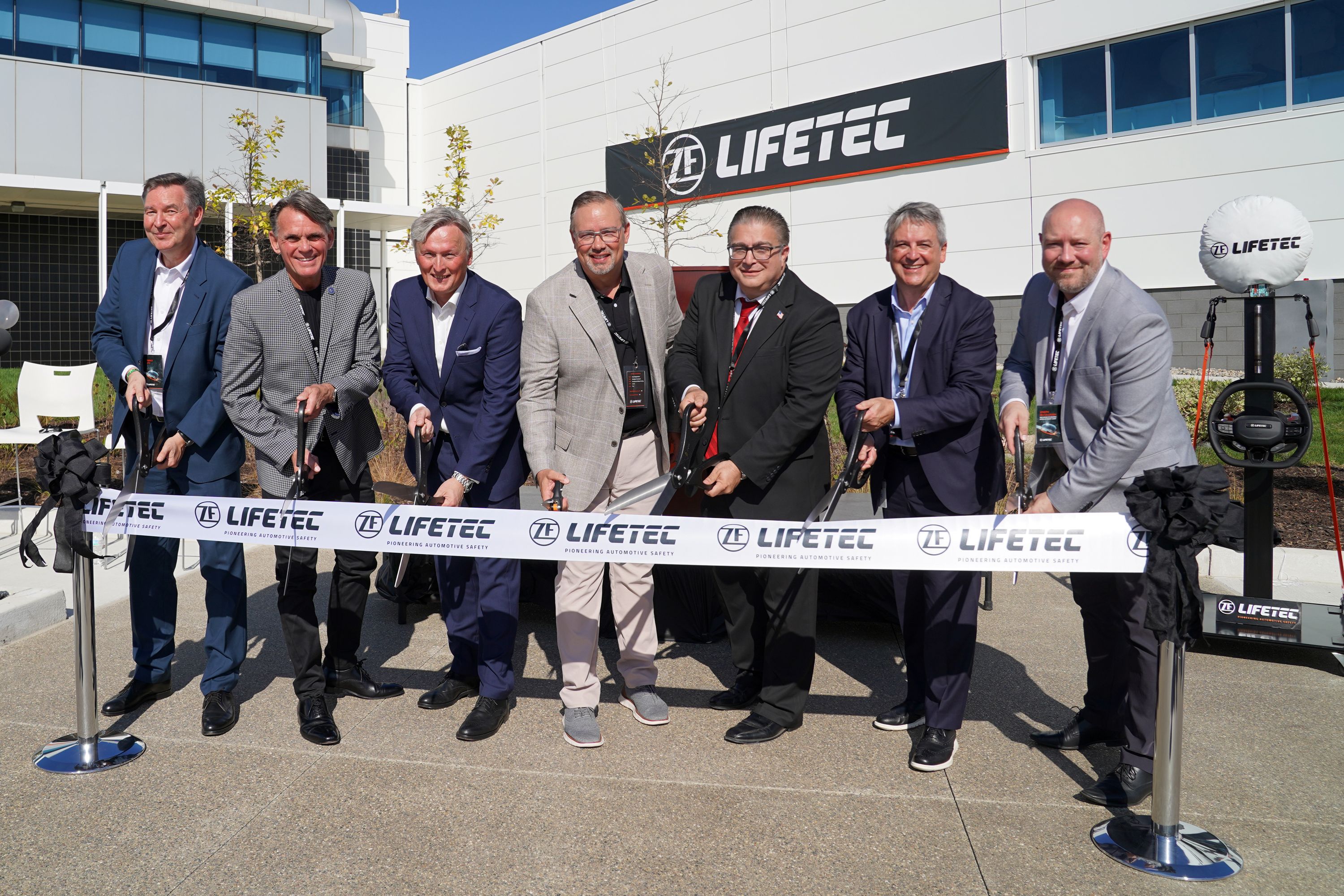 Eine Gruppe von sieben Männern in Geschäftskleidung durchschneidet während der großen Eröffnungsveranstaltung ein zeremonielles Band vor einem ZF Lifetec-Gebäude.