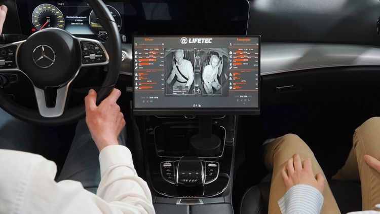 A driver and passenger sit inside a Mercedes-Benz vehicle while a ZF LIFETEC in-cabin monitoring system displays real-time safety and occupant data.