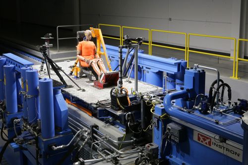 Crashtest-Dummy sitzt in einem Prüfstand, umgeben von blauen Hydraulikgeräten und Kameras in einer Laborumgebung.